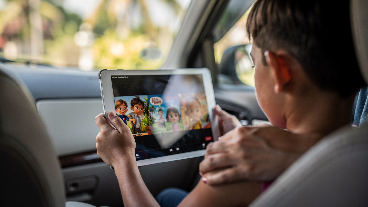 A child's hands hold an iPad showing a paused Disney+ movie, while the parent's hand gently rests on the child's shoulder, observing. The scene is set inside a comfortable, air-conditioned car in Bali, with glimpses of tropical scenery outside the window.