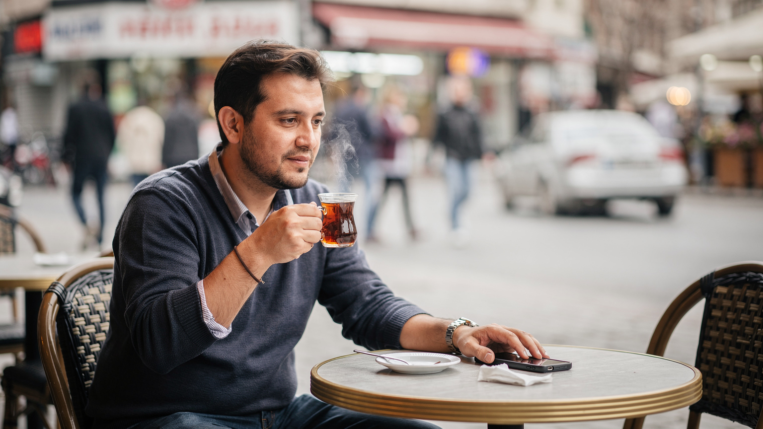 A person sits comfortably at an outdoor café table in Ankara's bustling Kızılay district, sipping Turkish tea from a traditional glass, and casually glancing at their smartphone, displaying a relaxed and connected travel experience.