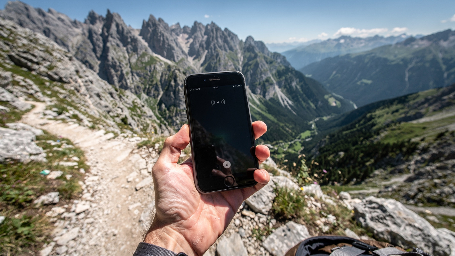 En mobiltelefon visar "Ingen service" medan den hålls upp av en person på en bergig vandringsled i norra Albanien, med vackra men avlägsna vyer i bakgrunden.