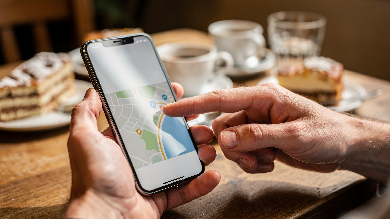 Close-up of a hand holding a smartphone, with another hand pointing at the screen, illustrating a moment of shared travel planning or troubleshooting. The setting is a cozy cafe in Budapest, Hungary.