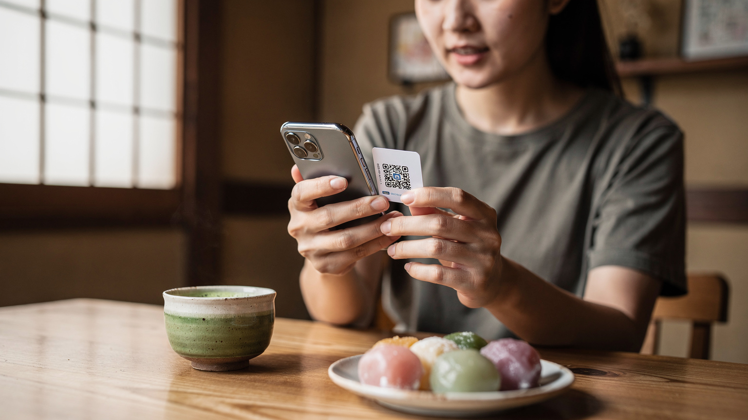 Một phụ nữ trẻ đang quét mã QR để kích hoạt eSIM trên điện thoại của mình, với một tách trà matcha và bánh mochi trên bàn cà phê trong một quán cà phê yên tĩnh ở khu phố Yanaka, Tokyo.