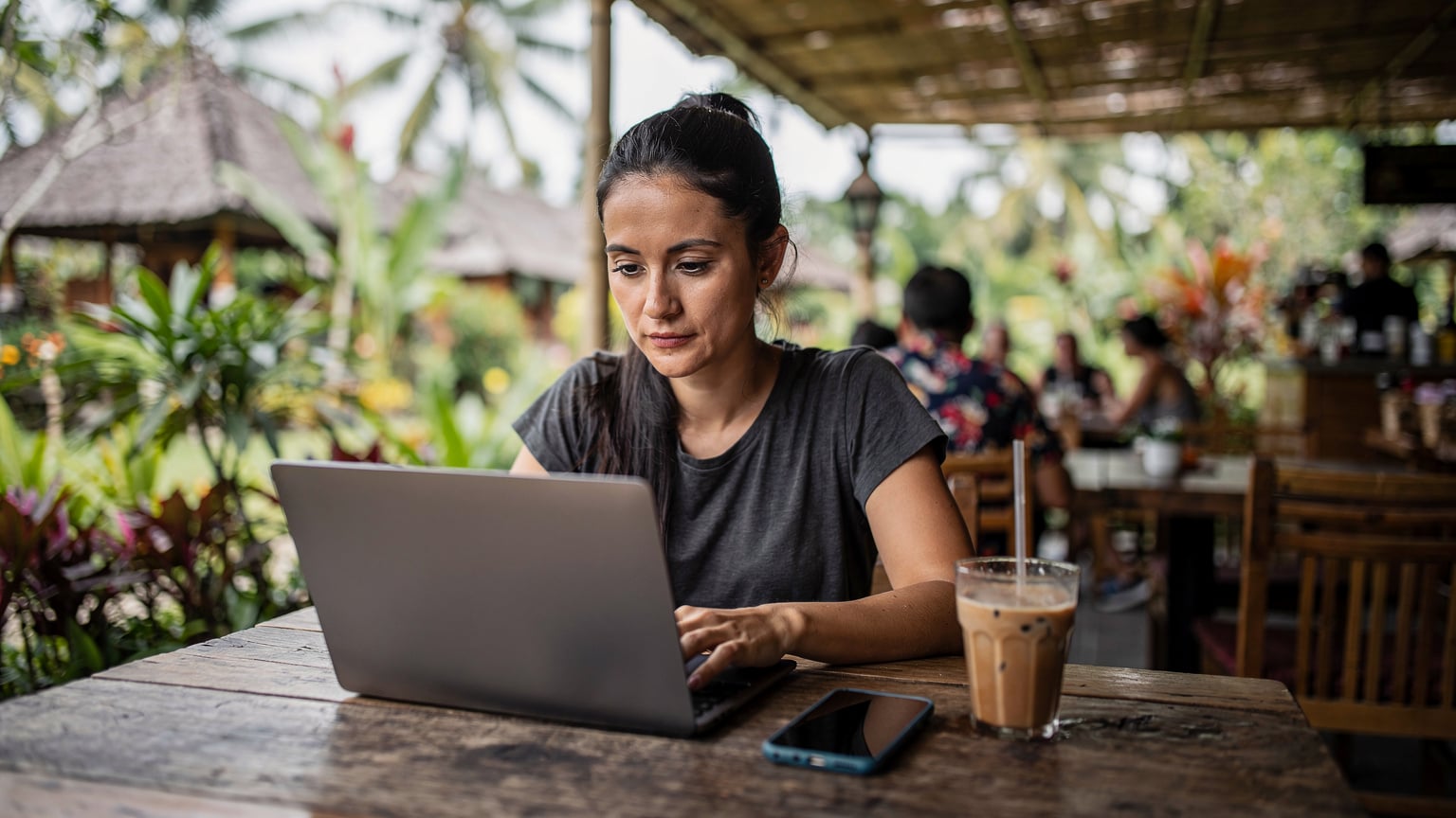 A woman with a laptop at an open-air cafe in Ubud, Bali, her phone displaying a Cellesim app next to a cup of coffee.