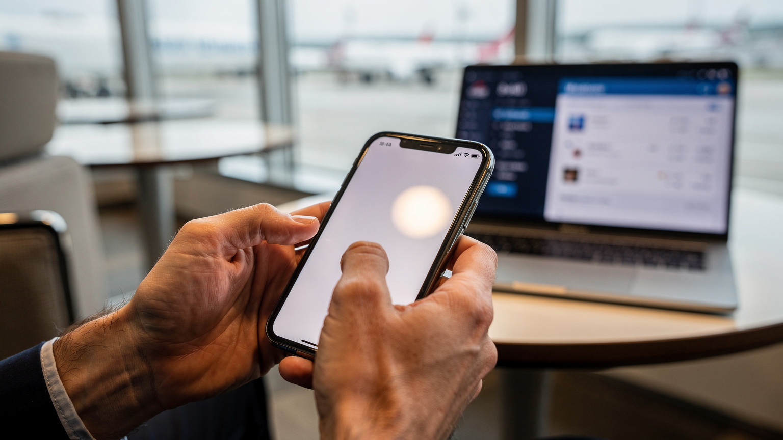 Close-up of a traveler's hands installing a Cellesim eSIM on a smartphone, with a laptop showing flight information to Indonesia in the background at an airport lounge.