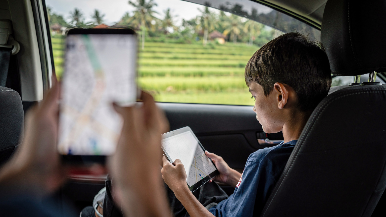 A child intently watching a downloaded movie on a tablet during a long car journey through the rice paddies of Bali, while a parent navigates on a smartphone in the foreground.