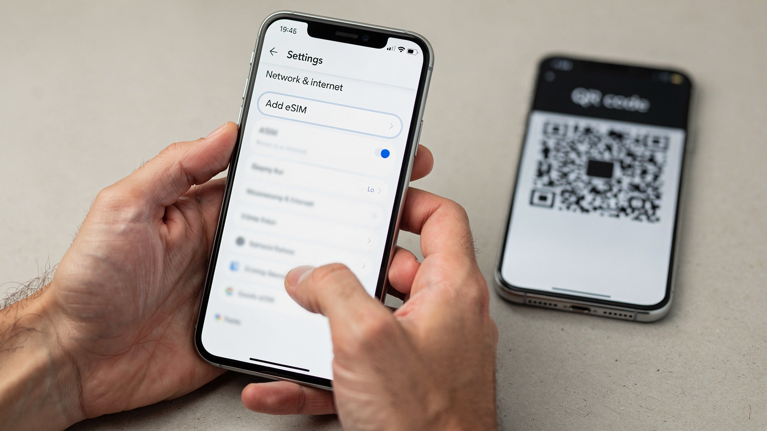 Un primo piano di mani che tengono uno smartphone Android, mostrando la schermata di impostazioni per l'aggiunta di una eSIM, con un codice QR sfocato in secondo piano su un altro dispositivo.