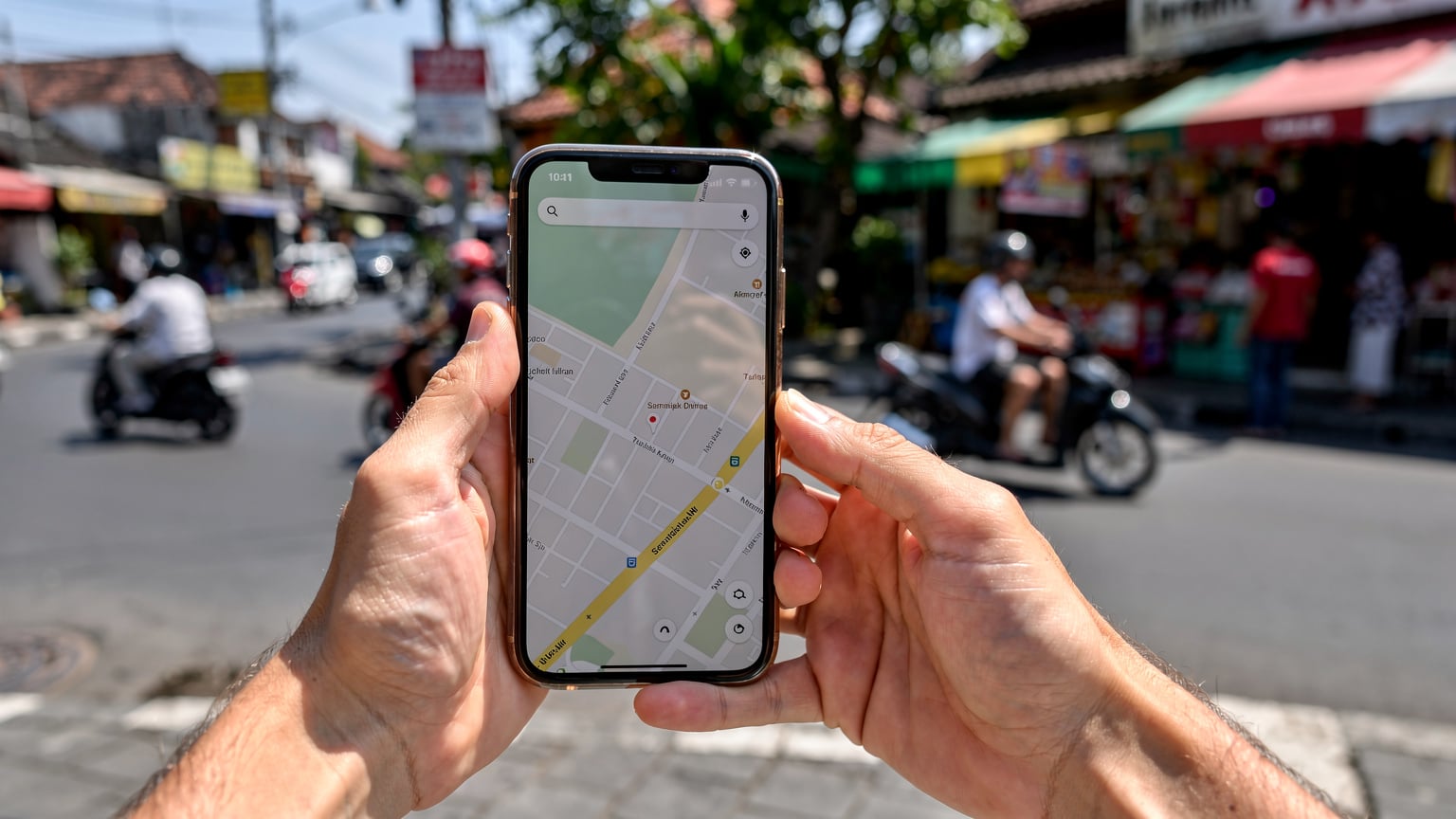 A person holds a smartphone displaying a map application, navigating a bustling street with traditional Indonesian architecture and other pedestrians in the background. The focus is on the phone and hands, with the street blurred slightly.