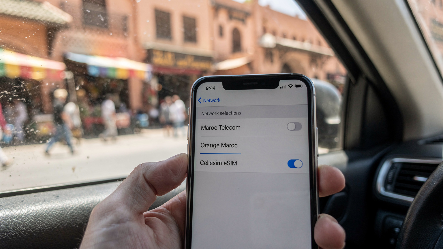 Ekran smartfona pokazujący dostępne sieci komórkowe w Maroku, z zaznaczoną opcją eSIM, na tle widoku z okna taksówki jadącej przez zatłoczoną ulicę Marrakeszu.