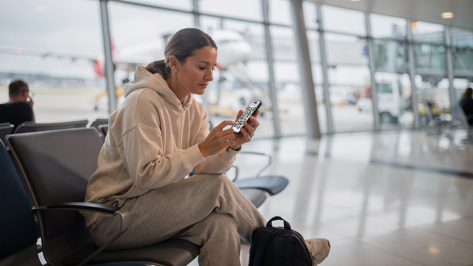Een vrouw scant een QR-code op haar smartphone om een eSIM te activeren, zittend in een drukke vertrekhal van Schiphol Airport met de vleugel van een vliegtuig op de achtergrond.