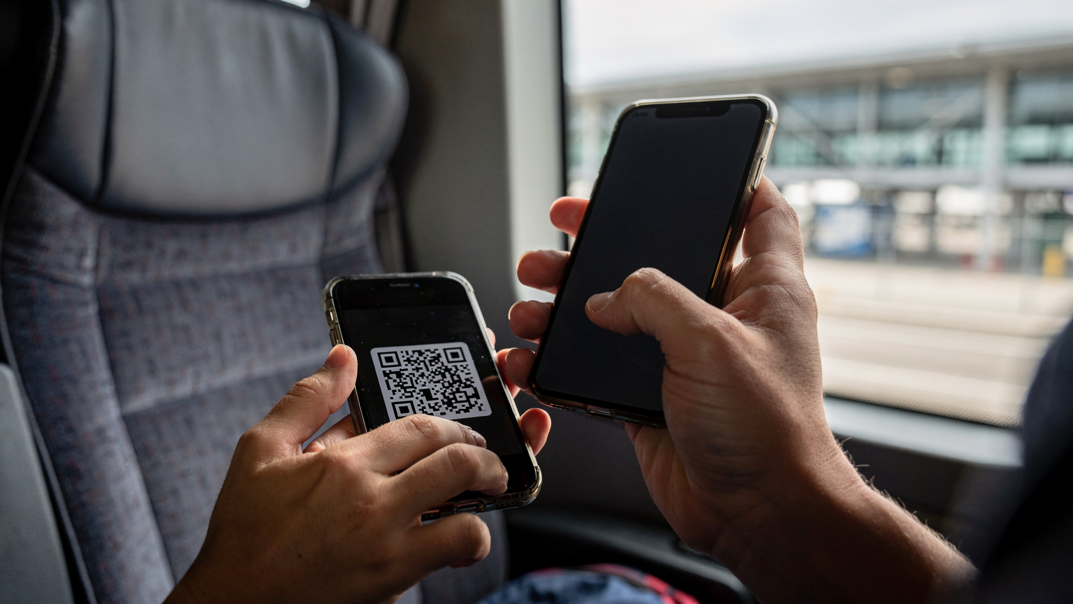 Zblízka na ruky mladého cestovateľa, ktorý drží smartfón a skenuje QR kód eSIM z druhého telefónu, pričom v pozadí je rozmazané okno autobusu a letiskový terminál.