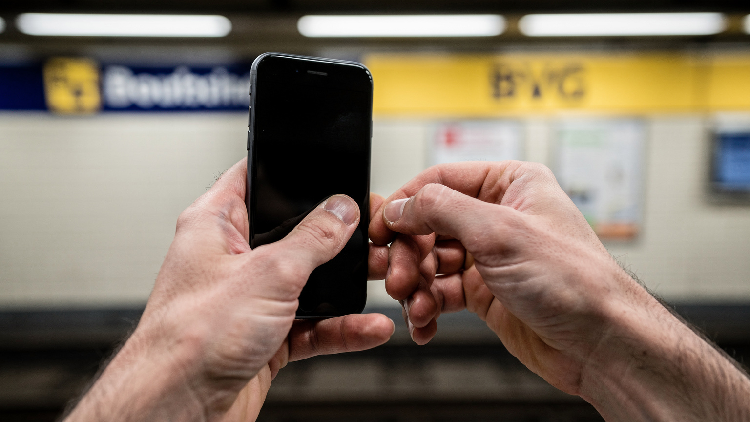 Un primo piano di mani che tengono uno smartphone, mentre un dito tocca lo schermo in un gesto di scorrimento verso l'alto, con lo sfondo sfocato di un cartello di una stazione della metropolitana di Berlino con il logo BVG.