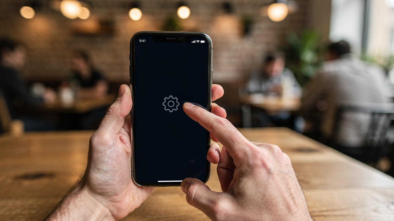 Nahaufnahme von Händen, die ein Smartphone halten, wobei der Finger auf das Symbol für die Einstellungen tippt, während der Hintergrund unscharf ein belebtes Café in Berlin zeigt.