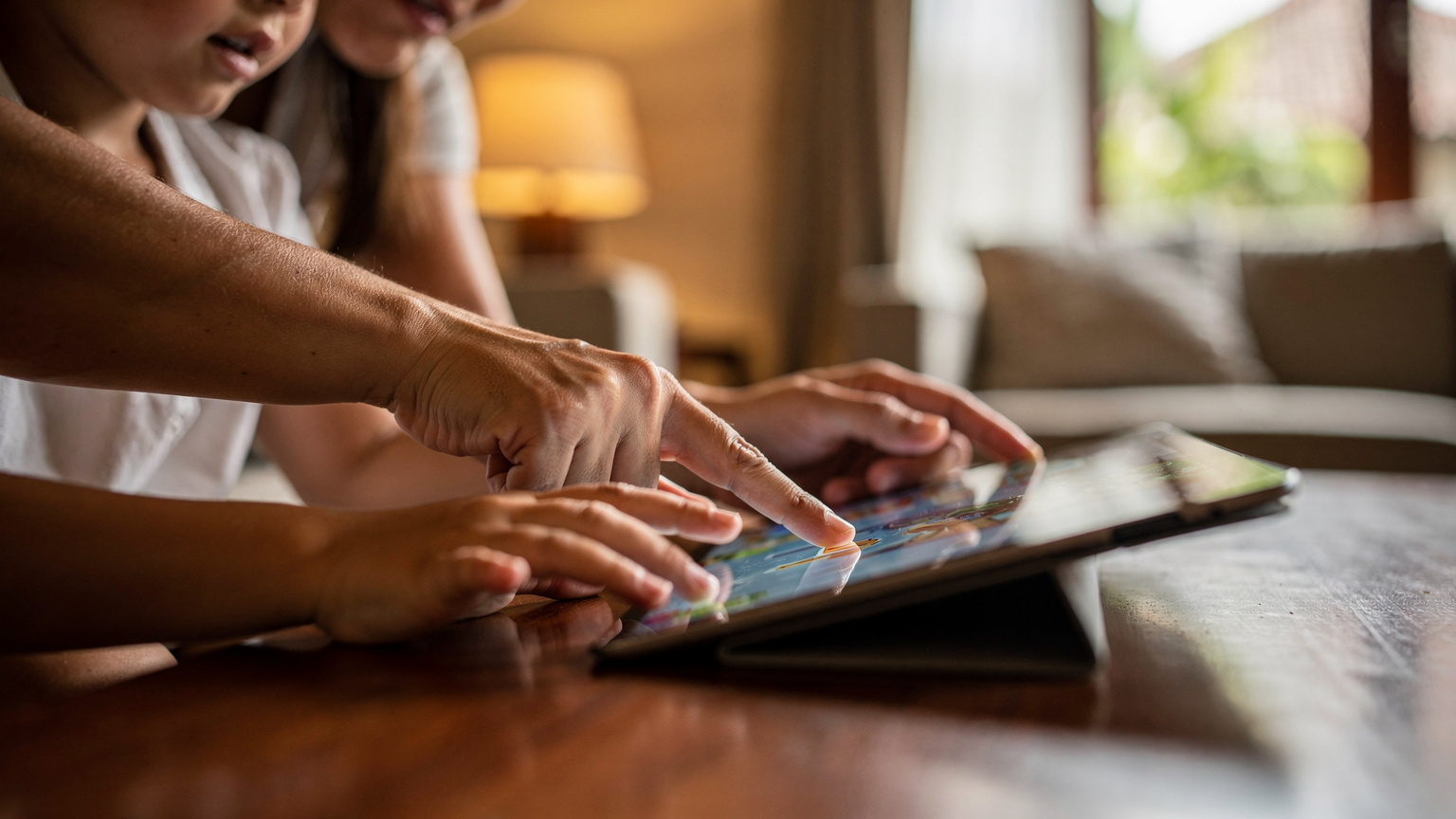 A mother gently guiding her child's hands on a tablet, showing them how to navigate a pre-downloaded game or educational app. The tablet screen shows a vibrant, child-friendly illustration without any readable text or UI.