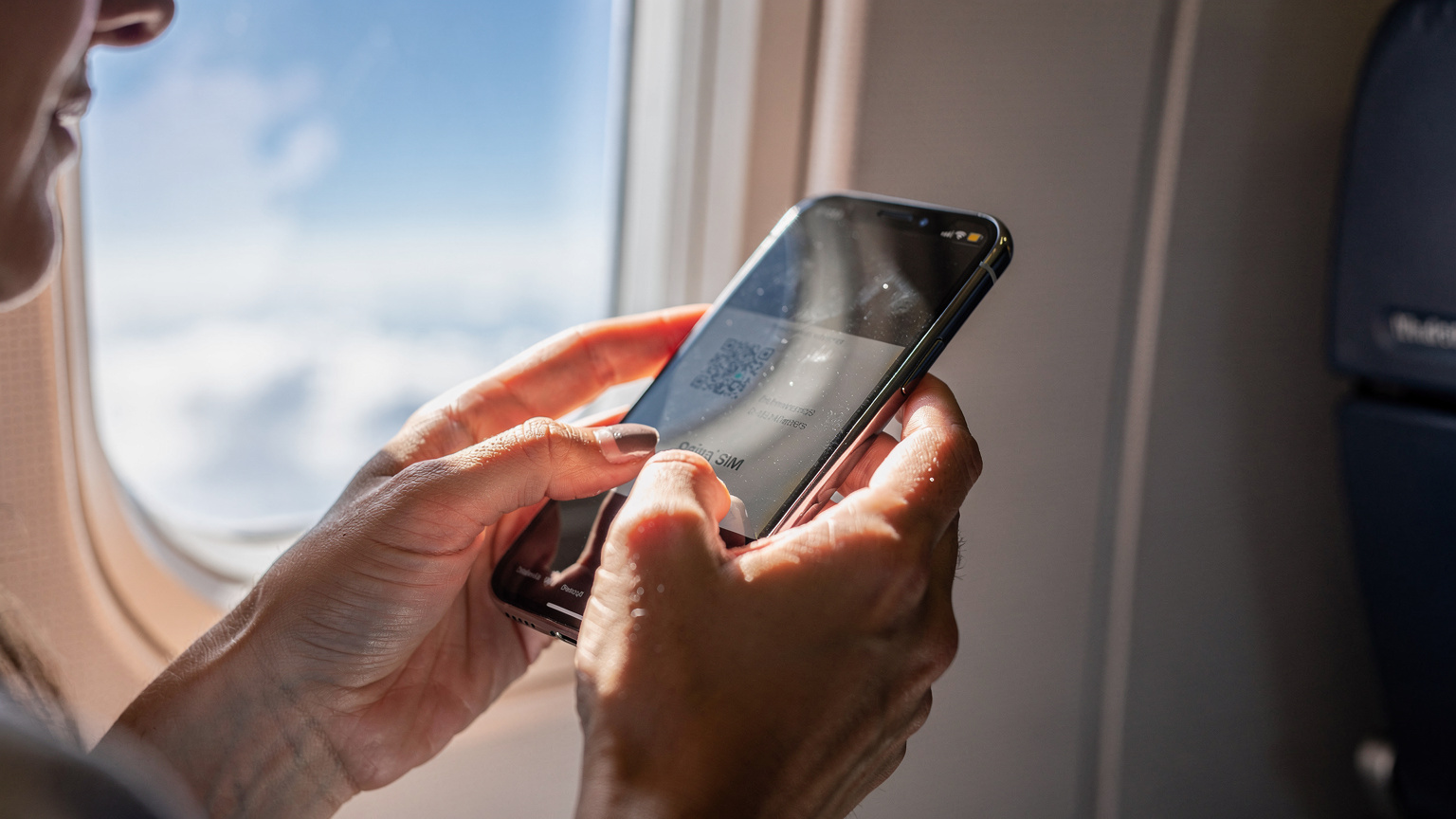 A close-up of a woman's hands holding an iPhone, scanning a QR code for eSIM activation, with the blurred background of an airplane window during a daytime flight.