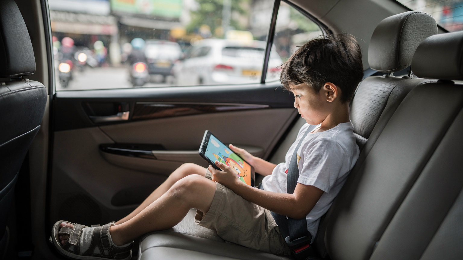 A child of about seven years old intently watching a cartoon on an iPad, sitting in the back seat of a moving taxi on a busy street in Jakarta.