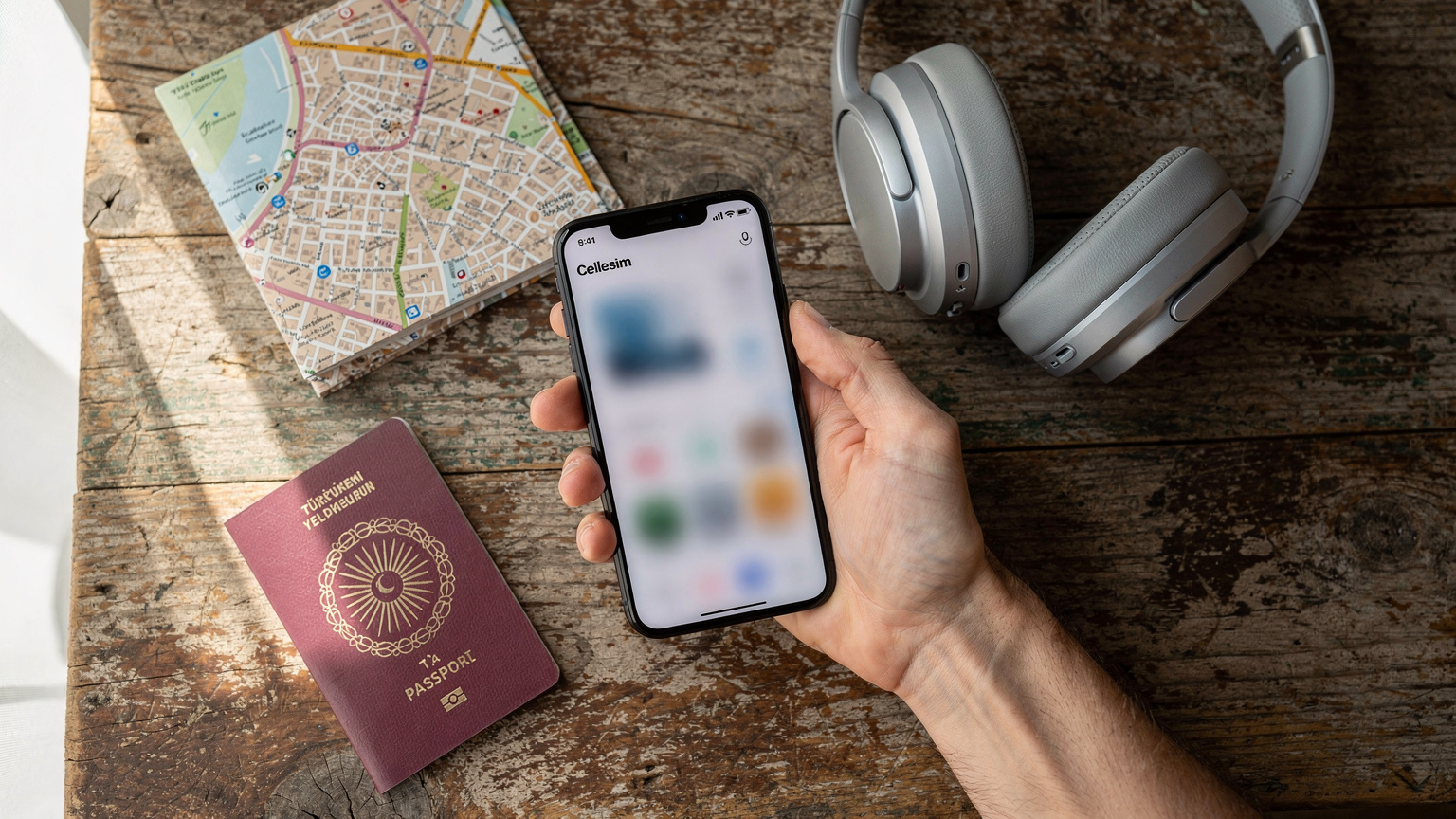 Bir seyyahın elinde, mobil cihazında Cellesim uygulamasının ana ekranı açıkken, arka planda pasaportu ve kulaklığı görülen bir düz üstü çekim.