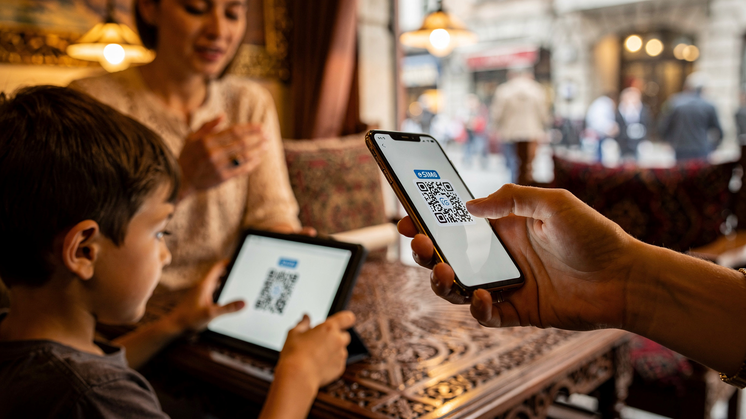 Bir anne, küçük oğlunun elindeki tablete eSIM QR kodunu taramak için kendi telefonunu kullanıyor. Arka planda otantik Türk kahve fincanları ve taze demlenmiş çay bardakları olan geleneksel bir Türk kafesi ortamı var.