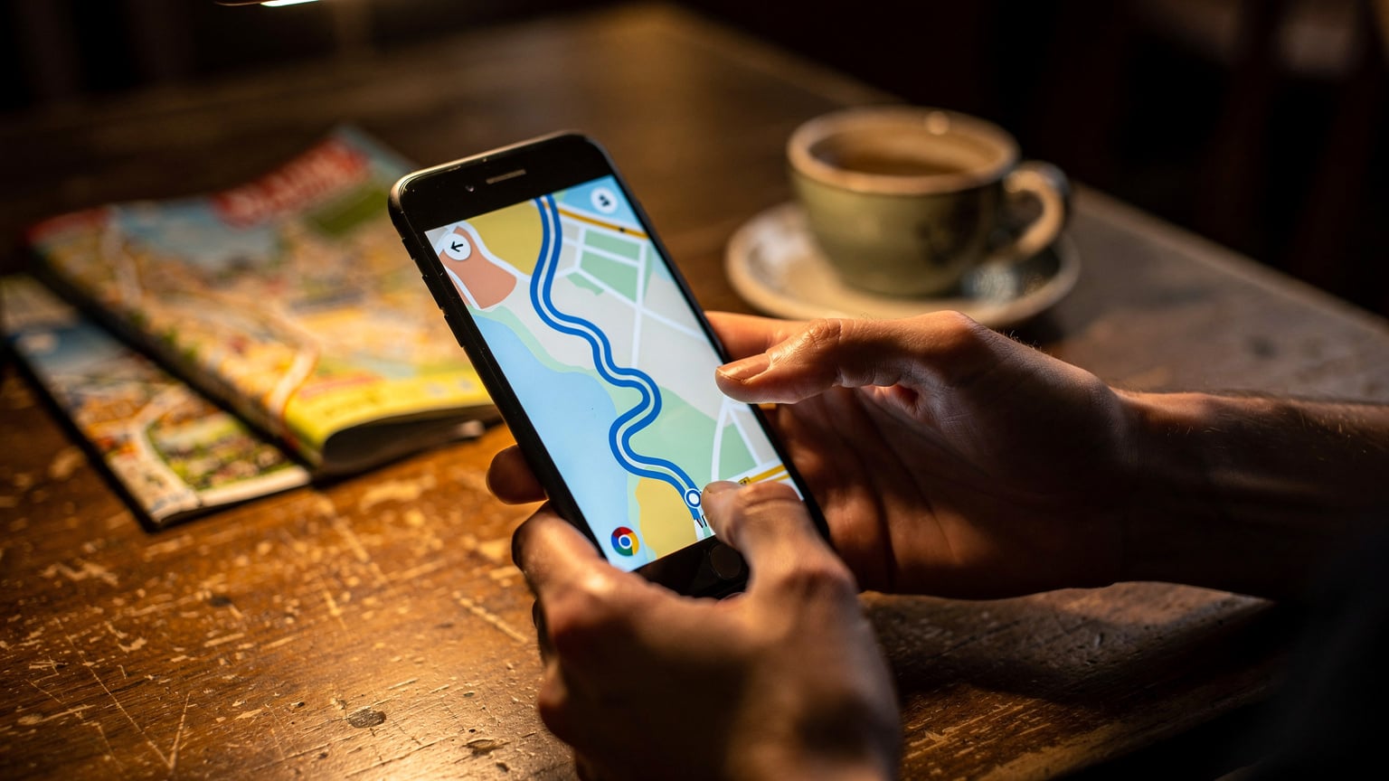 Eine Nahaufnahme von zwei Händen, die ein Smartphone halten. Der Daumen einer Hand wischt über den Bildschirm, auf dem eine farbige Karte mit einer blauen, markierten Route zu sehen ist. Im Vordergrund liegt unscharf ein zerknitterter Reiseführer mit Notizen.