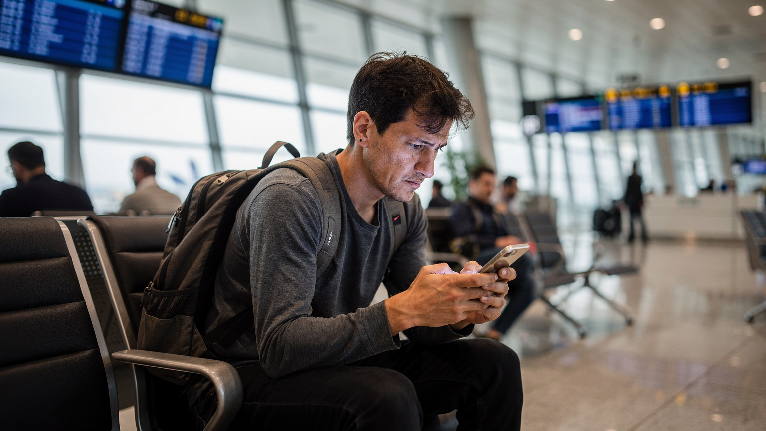 A traveler at an airport gate, looking at their phone screen with a slight frown, indicating a connectivity issue or confusion, while other passengers walk past in the background.