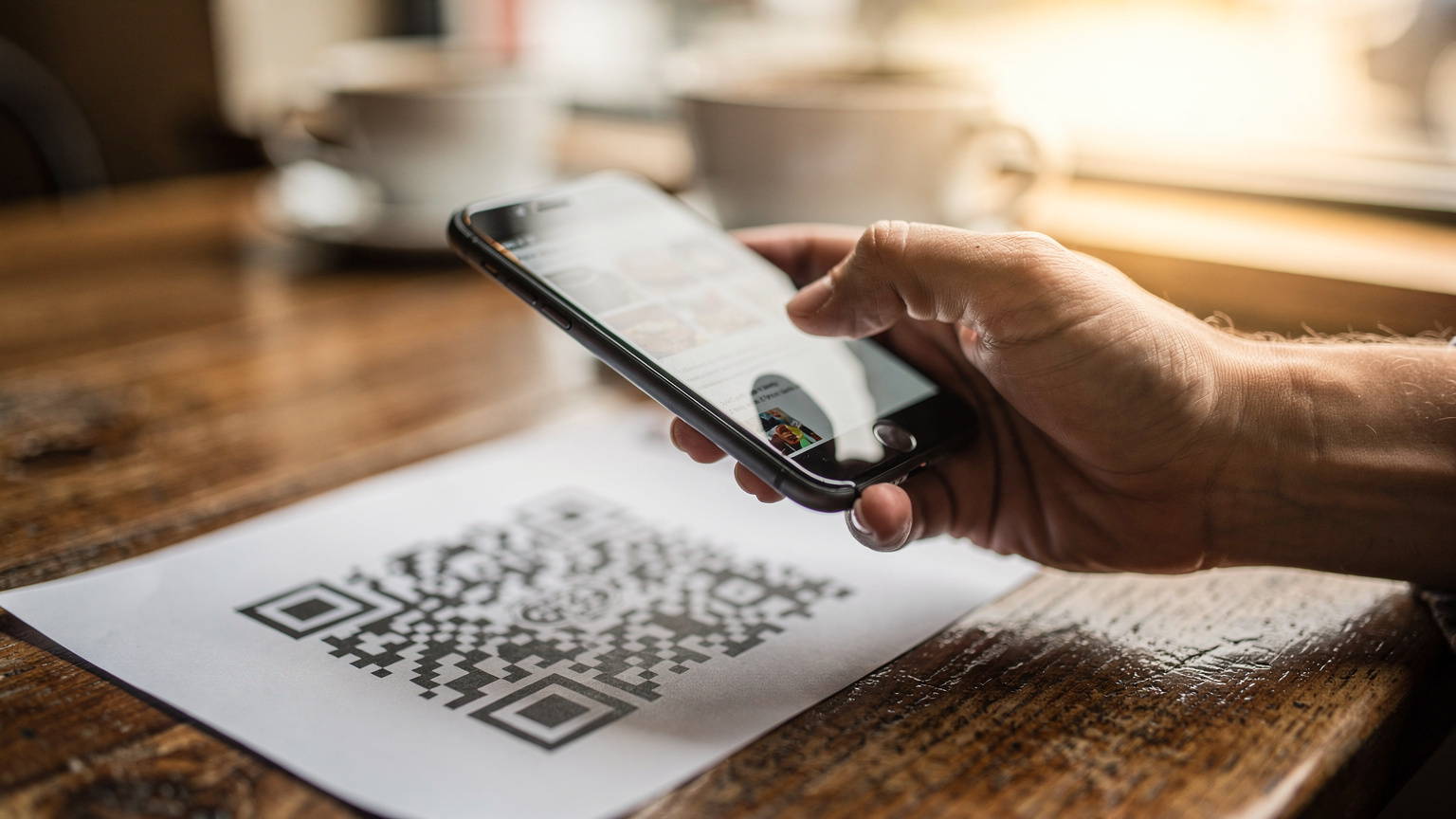 Main tenant un téléphone au-dessus d'un QR code imprimé sur une table de café, geste de scan