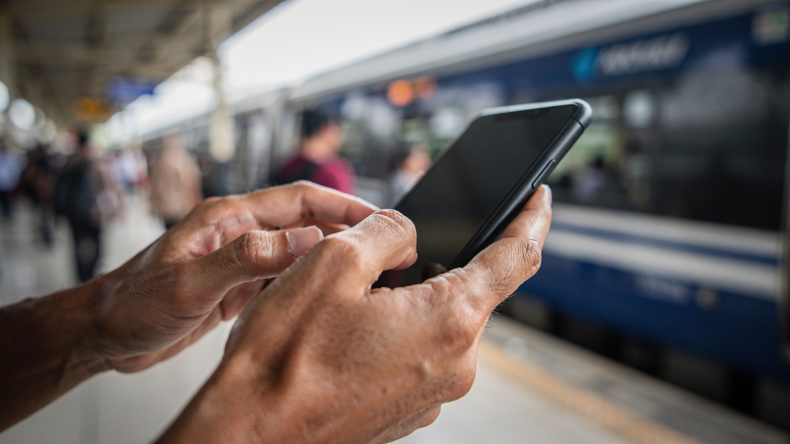 A close-up of a traveler's hands interacting with a phone at a train station in Bandung, Indonesia, with a subtle blur of people and trains in the background.