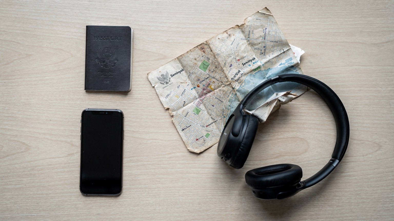 An editorial overhead flat-lay shot of a passport, a smartphone, noise-canceling headphones, and a crumpled paper map snippet of Seminyak, Bali, arranged on a light wooden table.