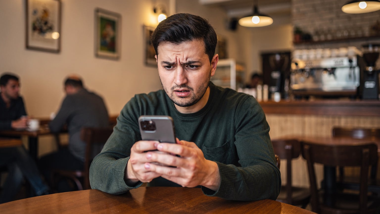 Bir kafeteryada oturan bir kişinin elinde telefonla sorun yaşadığı, kaşlarını çattığı ve ekrana dikkatle baktığı bir yakın çekim.