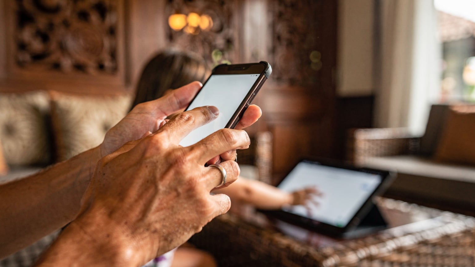 Close-up of a parent's hands configuring screen time settings on an iPhone, with a blurred child's hand reaching for an iPad in the background, set in a cozy hotel lobby in Ubud, Bali.
