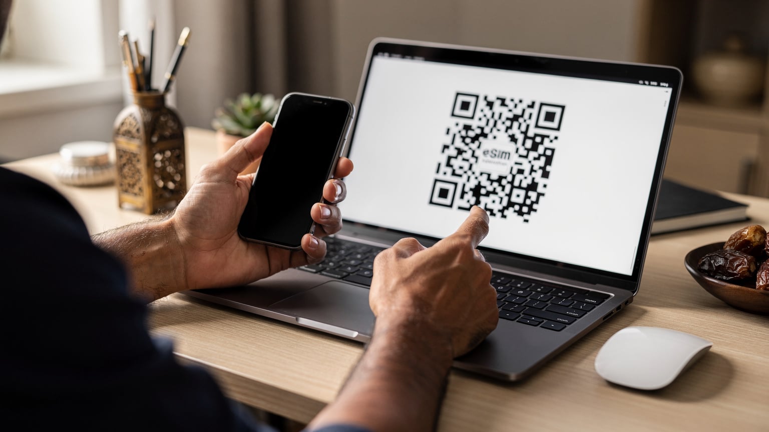 يد رجل عربي تحمل هاتفًا ذكيًا وتوجهه نحو شاشة كمبيوتر محمول تعرض رمز QR، في مكتب منزلي مضاء جيدًا ومجهز بأدوات مكتبية بسيطة.