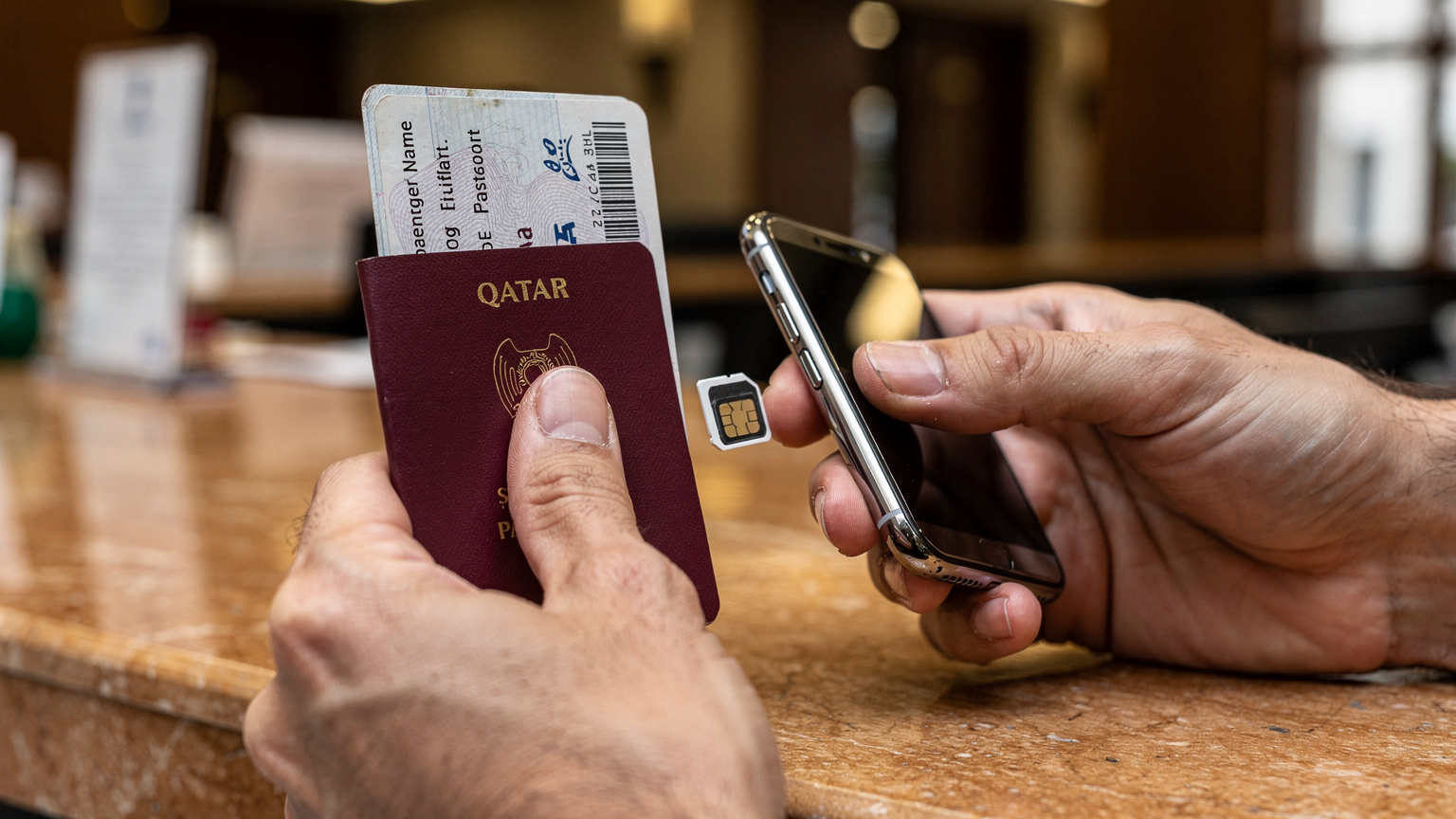 يدا مسافر تحملان وثائق السفر (جواز سفر قطري) وشريحة SIM بلاستيكية جديدة بينما يحاول إدخالها في هاتف ذكي، مع خلفية غير واضحة لمكتب استقبال في فندق في منطقة السلطان أحمد.