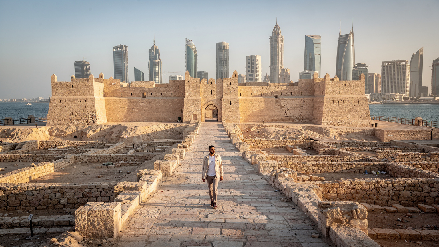 منظر علوي بانورامي لقلعة البحرين الأثرية مع خليج المنامة في الخلفية، وشخص يمشي على الممشى الأثري.