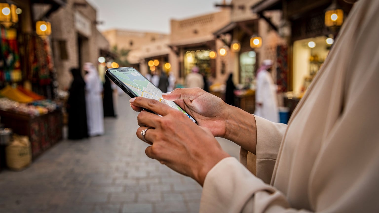 يد امرأة عربية تحمل هاتفاً ذكياً وتستخدم خرائط جوجل لتحديد الاتجاهات في سوق واقف بالدوحة، مع تفاعل عائلي في الخلفية، تعكس أجواء رمضان الهادئة.