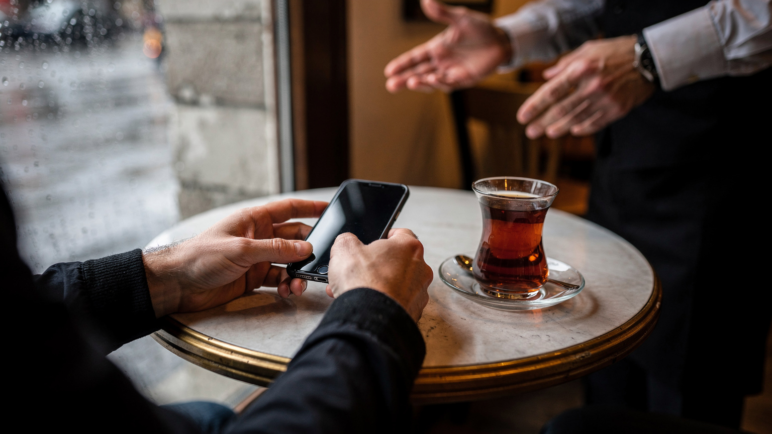 Viaggiatore con telefono in mano al tavolino di un caffè a Karaköy, Istanbul, con un bicchiere di tè turco davanti