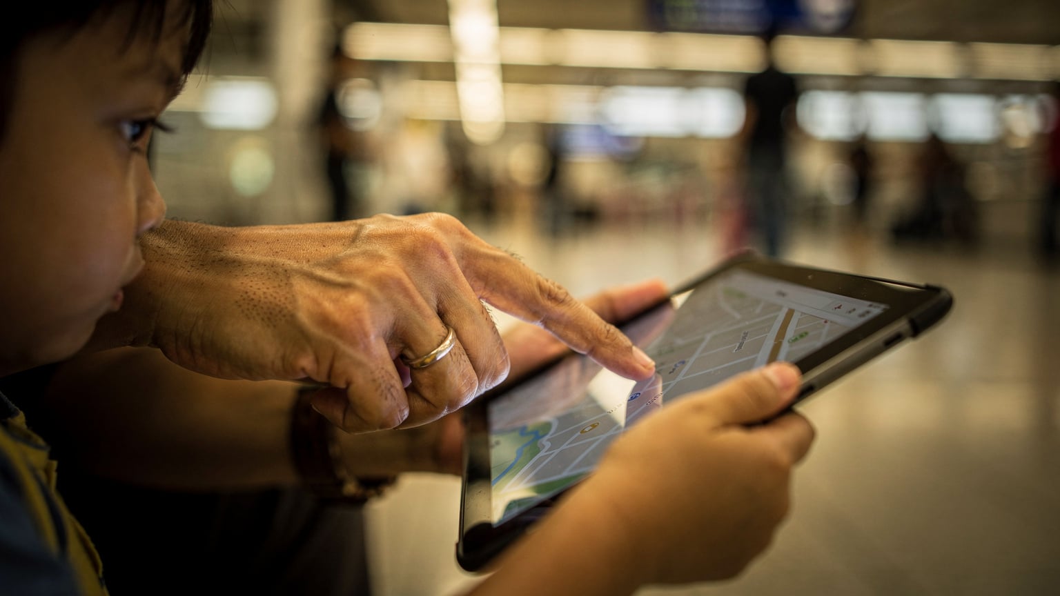 A parent's hands gently guiding a child's finger on a tablet screen, showing them how to zoom in on a map application while waiting at a bustling transit hub in Jakarta.