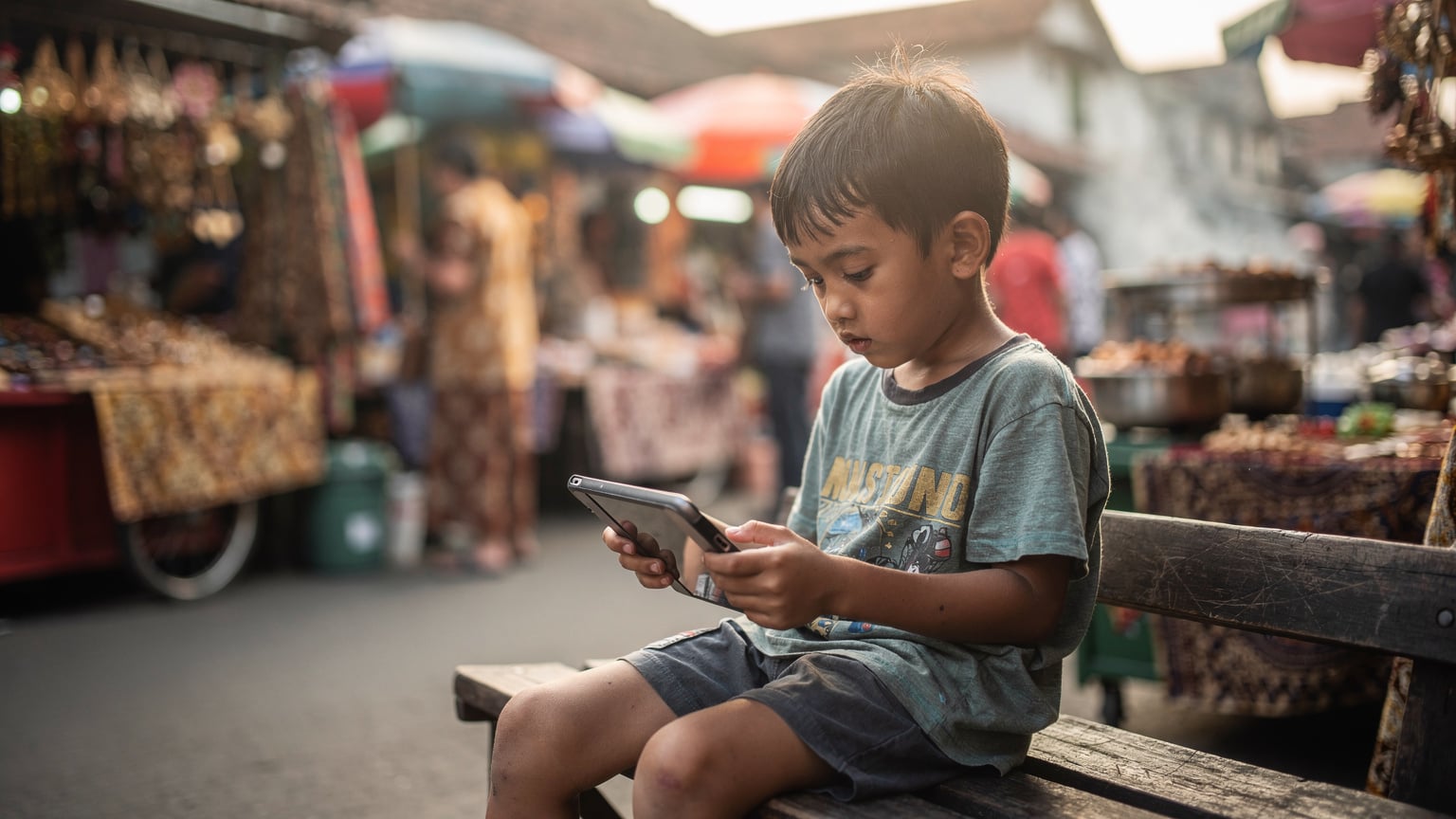 A child engrossed in a tablet game while sitting on a bench in the bustling streets of Yogyakarta, Indonesia, oblivious to the vibrant market activity around them.