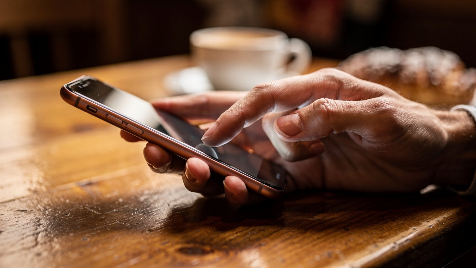 Nahaufnahme der Hände eines Reisenden, der ein Smartphone hält und mit dem Daumen über den Bildschirm wischt, während er an einem Holztisch in einem gemütlichen Café in Salzburg sitzt.