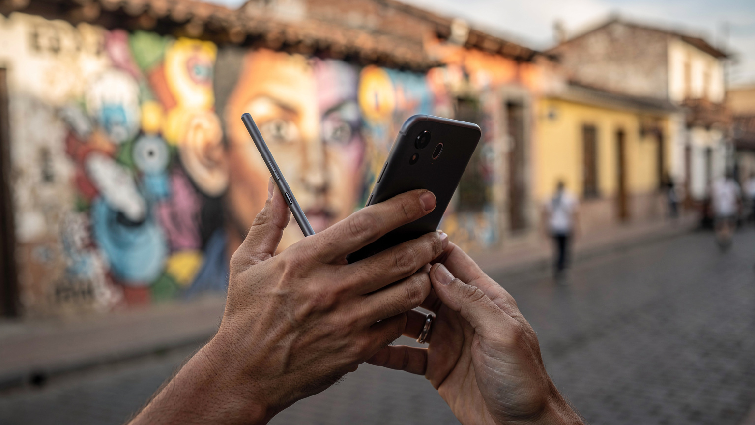 Close-up on a pair of hands holding a smartphone, with a blurred background of a street art mural in the La Candelaria district of Bogotá, Colombia.