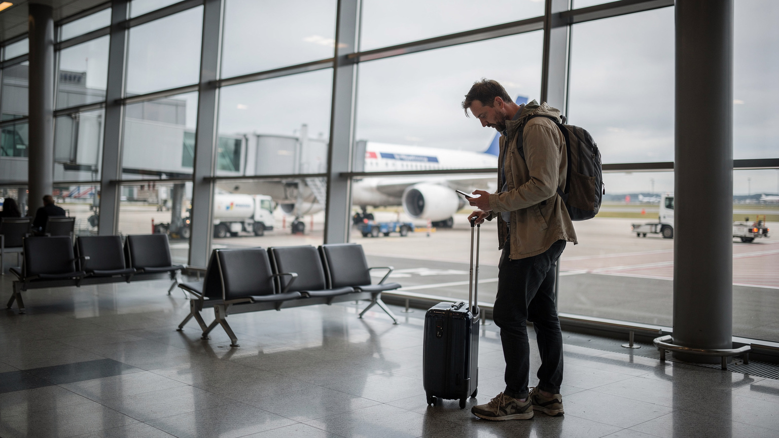 Ein Reisender am Flughafen Berlin Brandenburg (BER) überprüft nervös sein Smartphone am Gate, während im Hintergrund ein Flugzeug betankt wird.