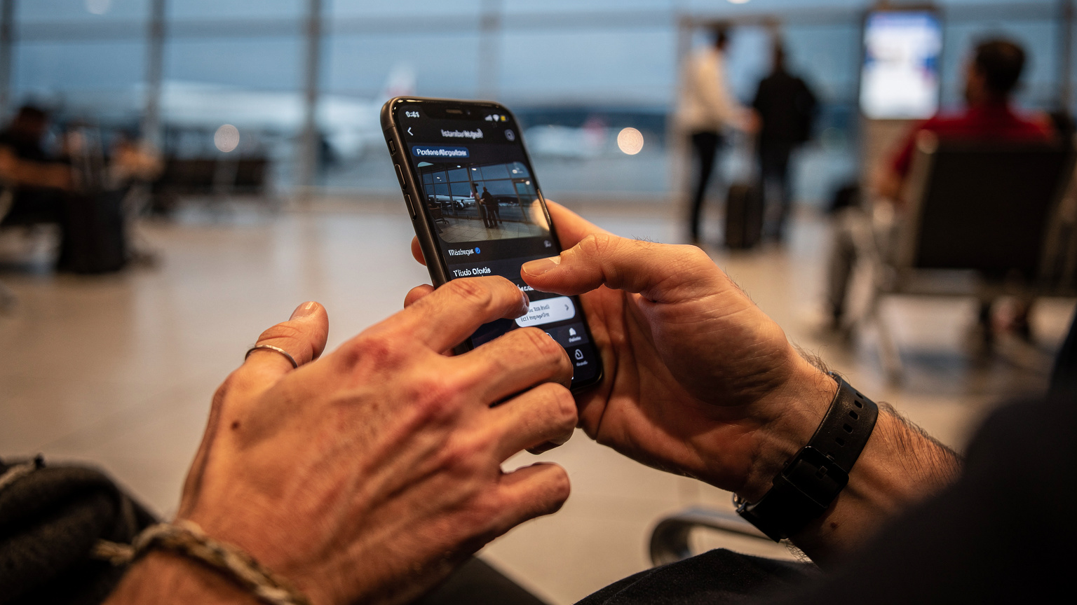 İstanbul Havalimanı dış hatlar terminalinde, kalkış saatini bekleyen bir yolcu. Telefonunu tutan elleri ekranda hızlıca işlem yapıyor, ancak ekran içeriği bulanık.