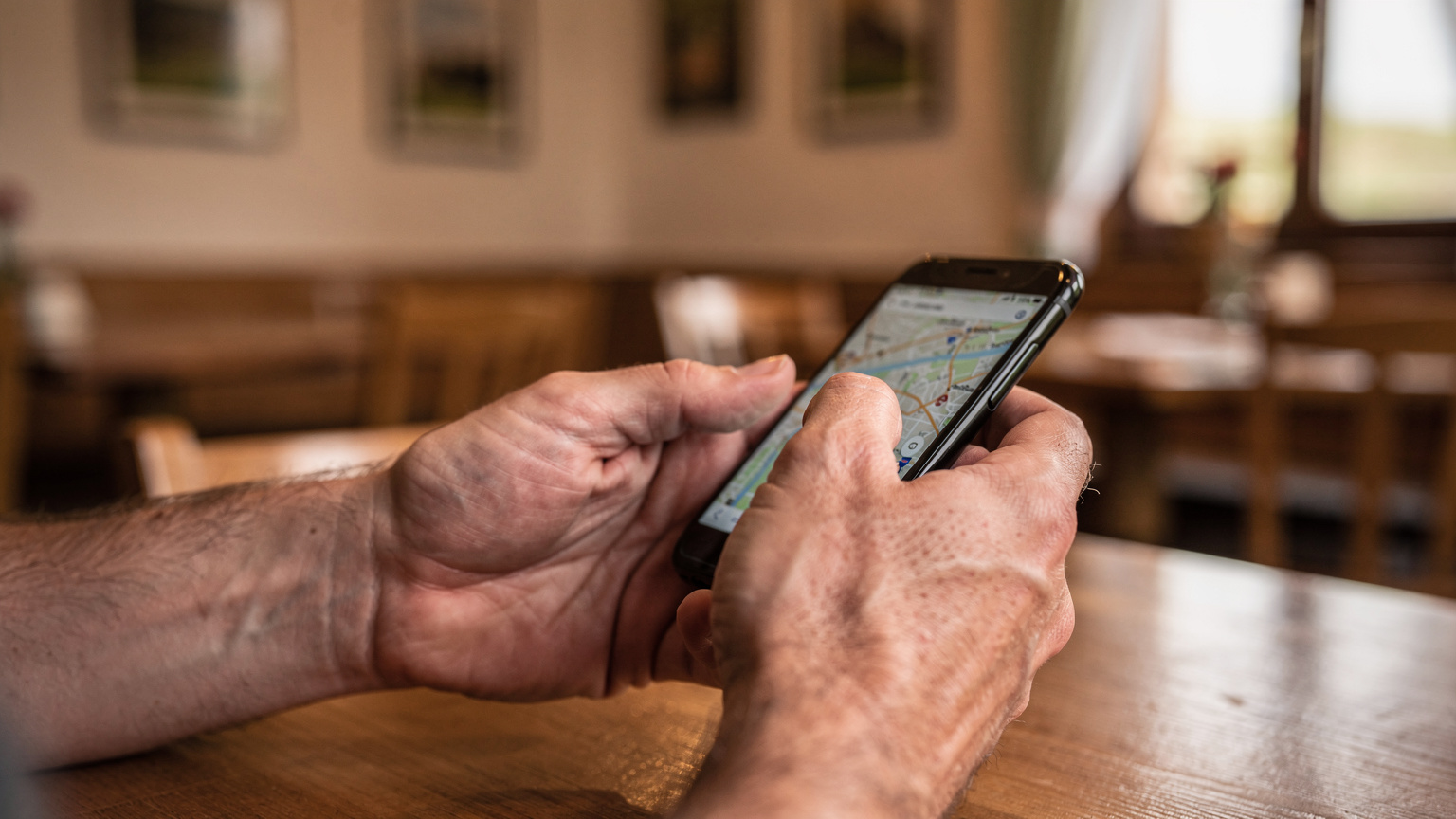 Nahaufnahme von Händen, die ein Smartphone halten, während ein Daumen über eine Karte wischt, um eine alternative Route zu finden, mit einem unscharfen Bild eines Straßenrestaurants im Hintergrund.