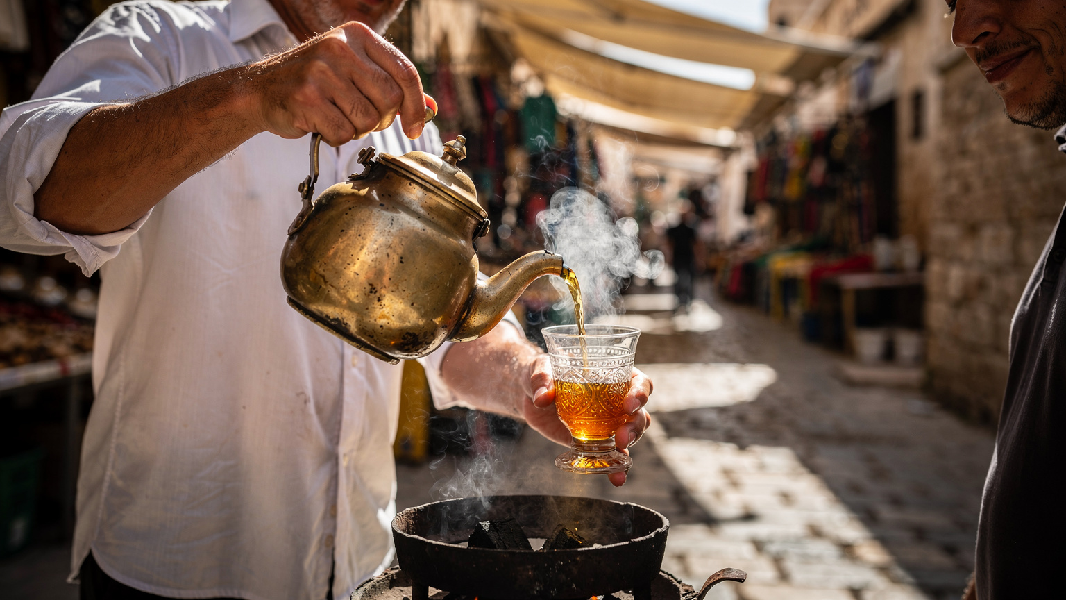 Un vendeur de thé à la menthe dans la médina de Sousse, brasero fumant et verres dorés, un client attend en souriant