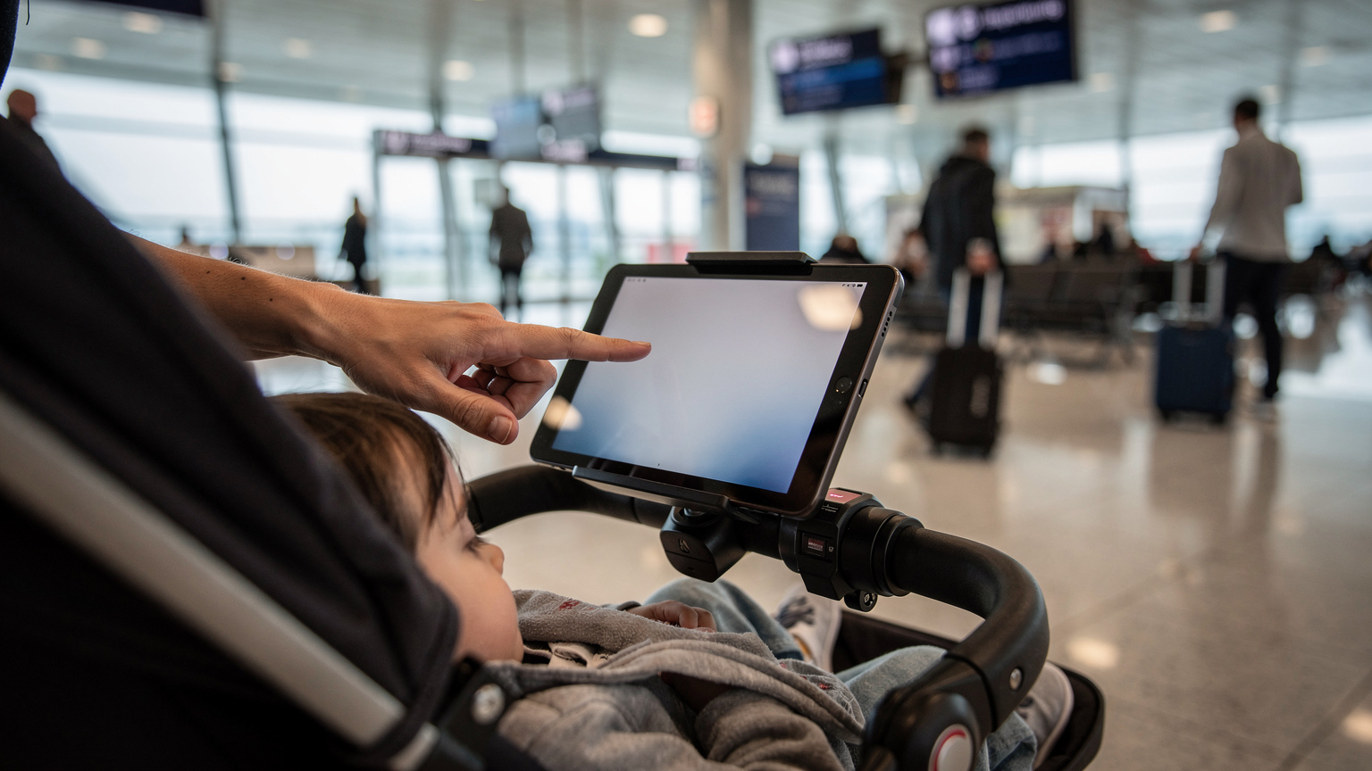 İstanbul Havalimanı'nda, bir annenin eli çocuk arabasındaki uyuyan çocuğunun tablet ekranında Wi-Fi bağlantı durumunu kontrol ediyor. Arka planda hareketli terminalin genel manzarası var, insanlar geçiyor ve ışıklar parlıyor.