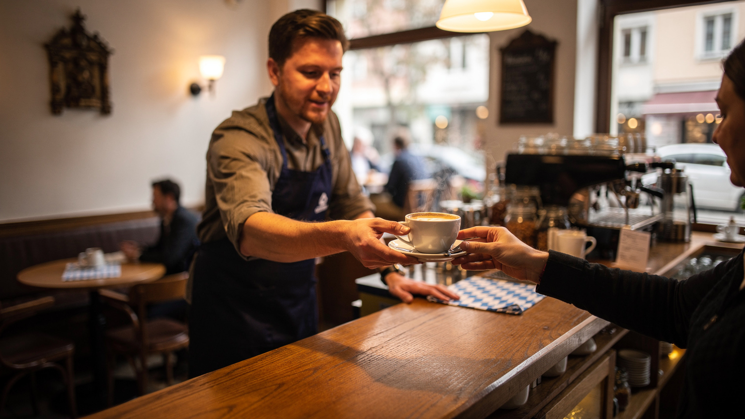 Ein Barista in einem Münchener Café reicht einer Hand eine Tasse Kaffee über die Theke, im Hintergrund sind angedeutet weitere Gäste und gedämpftes Licht.