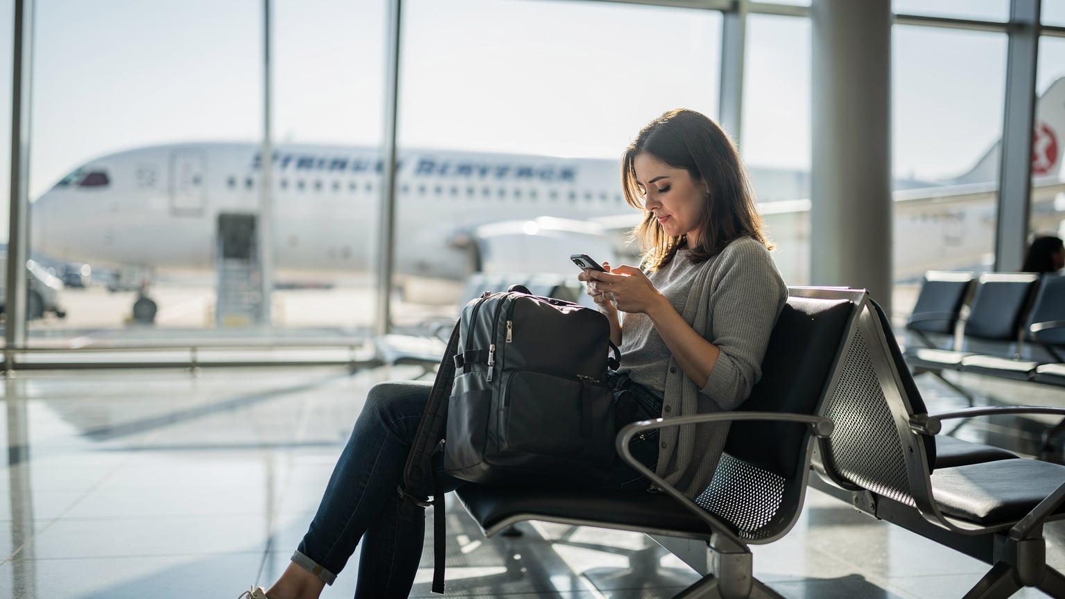 Woman at Istanbul Airport gate, focused on her smartphone, with a plane visible outside.