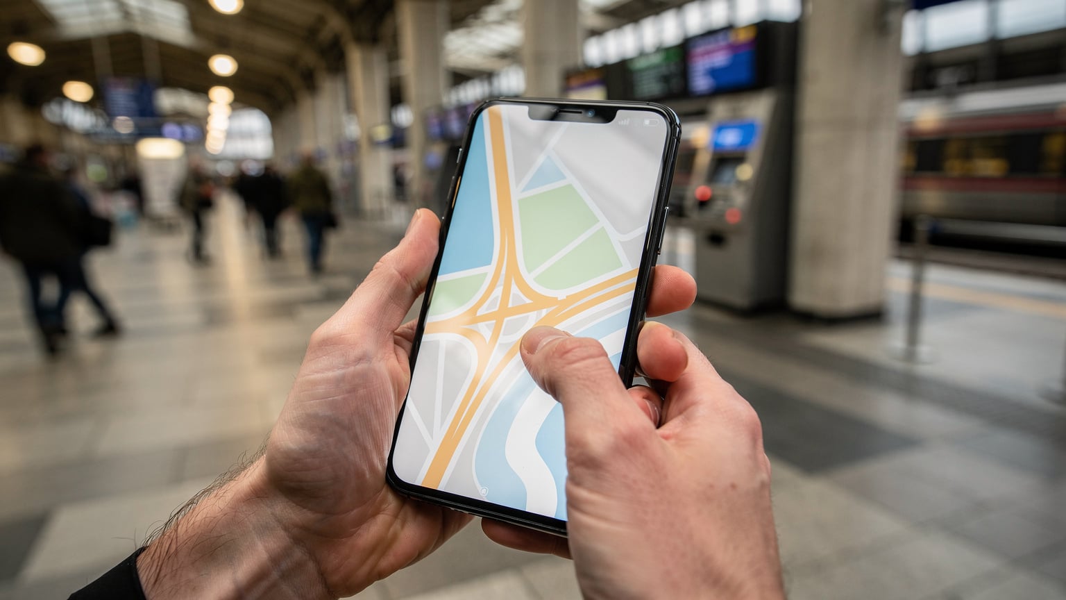 Nahaufnahme von Händen, die ein Smartphone halten, wobei der Fokus auf den Daumen liegt, der über den Bildschirm wischt. Das Display zeigt eine Karte oder eine Reise-App, ohne dass Texte oder Logos sichtbar sind. Der Hintergrund ist leicht verschwommen, deutet aber auf eine geschäftige Umgebung in einer europäischen Stadt hin.