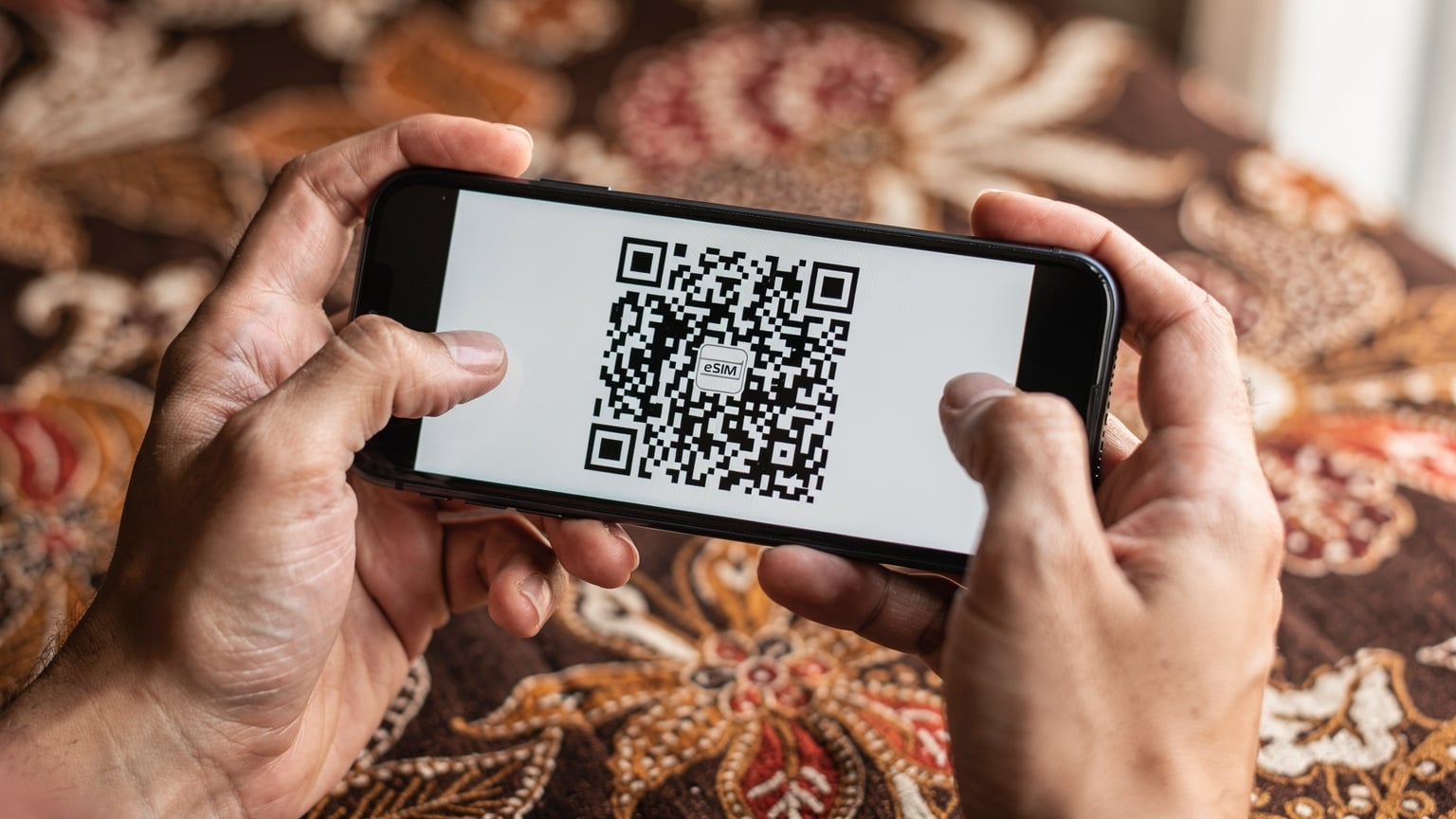 A close-up shot of a pair of human hands holding a smartphone, with the screen displaying a QR code. The hands are slightly blurred, emphasizing the QR code, against a backdrop of a blurred traditional Indonesian batik fabric pattern.