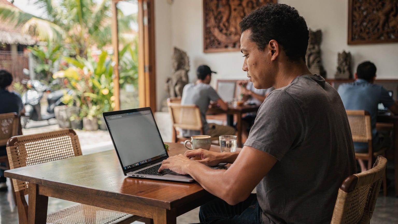 A digital nomad works diligently on a laptop at a rustic wooden table in a vibrant, open-air cafe in Canggu, Bali. Sunlight streams through lush tropical plants in the background. A ceramic mug of coffee and a small notebook are on the table next to the laptop, and the person's hands are actively typing.