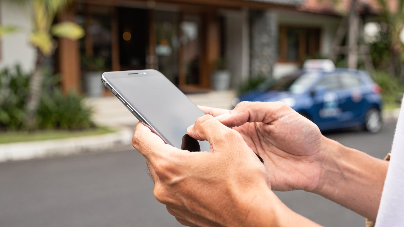 Close-up of a traveler's hands holding a smartphone, with fingers navigating settings on the screen, while standing outside a hotel in Seminyak, Bali, waiting for a taxi.