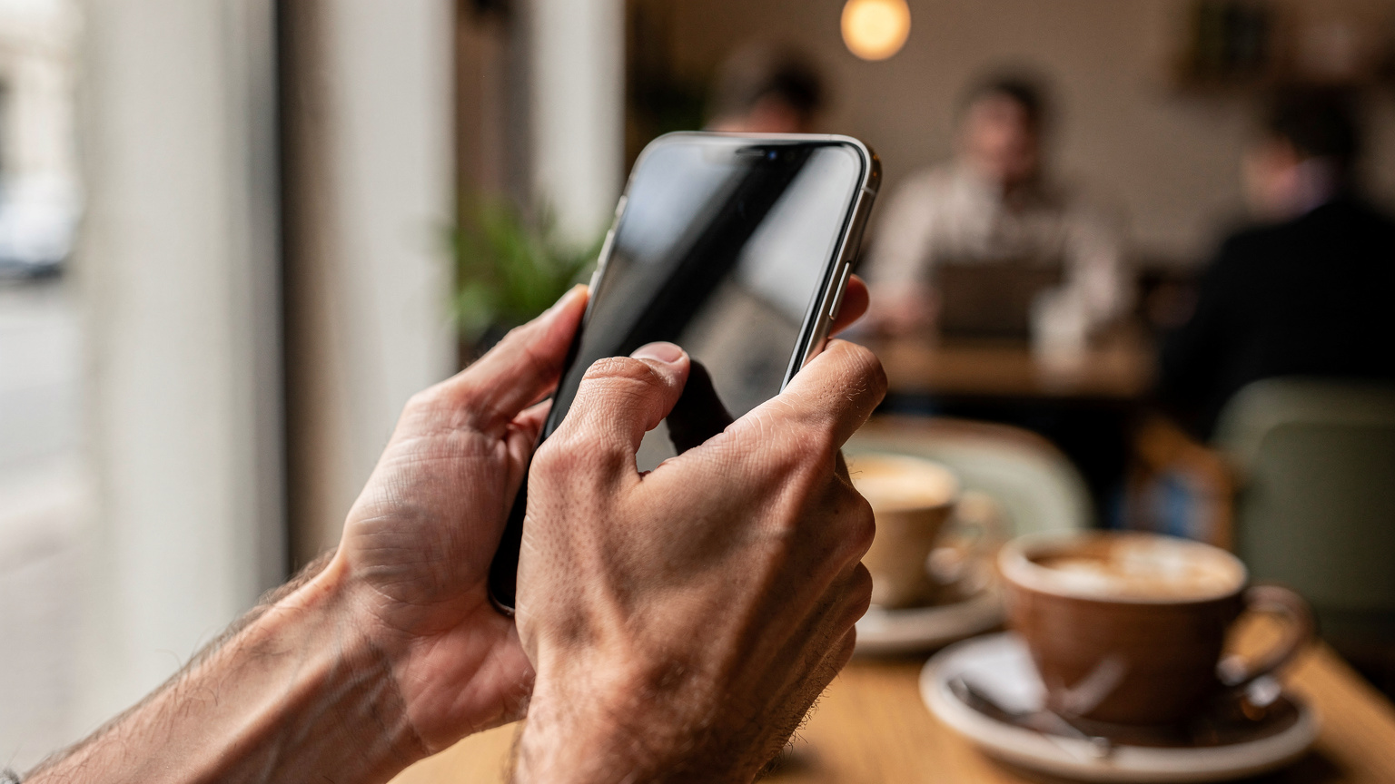 Nahaufnahme von Händen, die ein Smartphone halten und über den Bildschirm wischen, um eine eSIM einzurichten, mit verschwommenem Hintergrund eines Berliner Cafés.