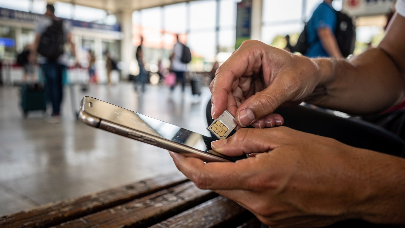 Nahaufnahme der Hände eines Reisenden, der versucht, eine winzige SIM-Karte in ein Smartphone einzulegen, während er auf einer Holzbank in einem belebten Busbahnhof in Tirana, Albanien, sitzt, im Hintergrund verschwimmen Reisende und Gepäck.