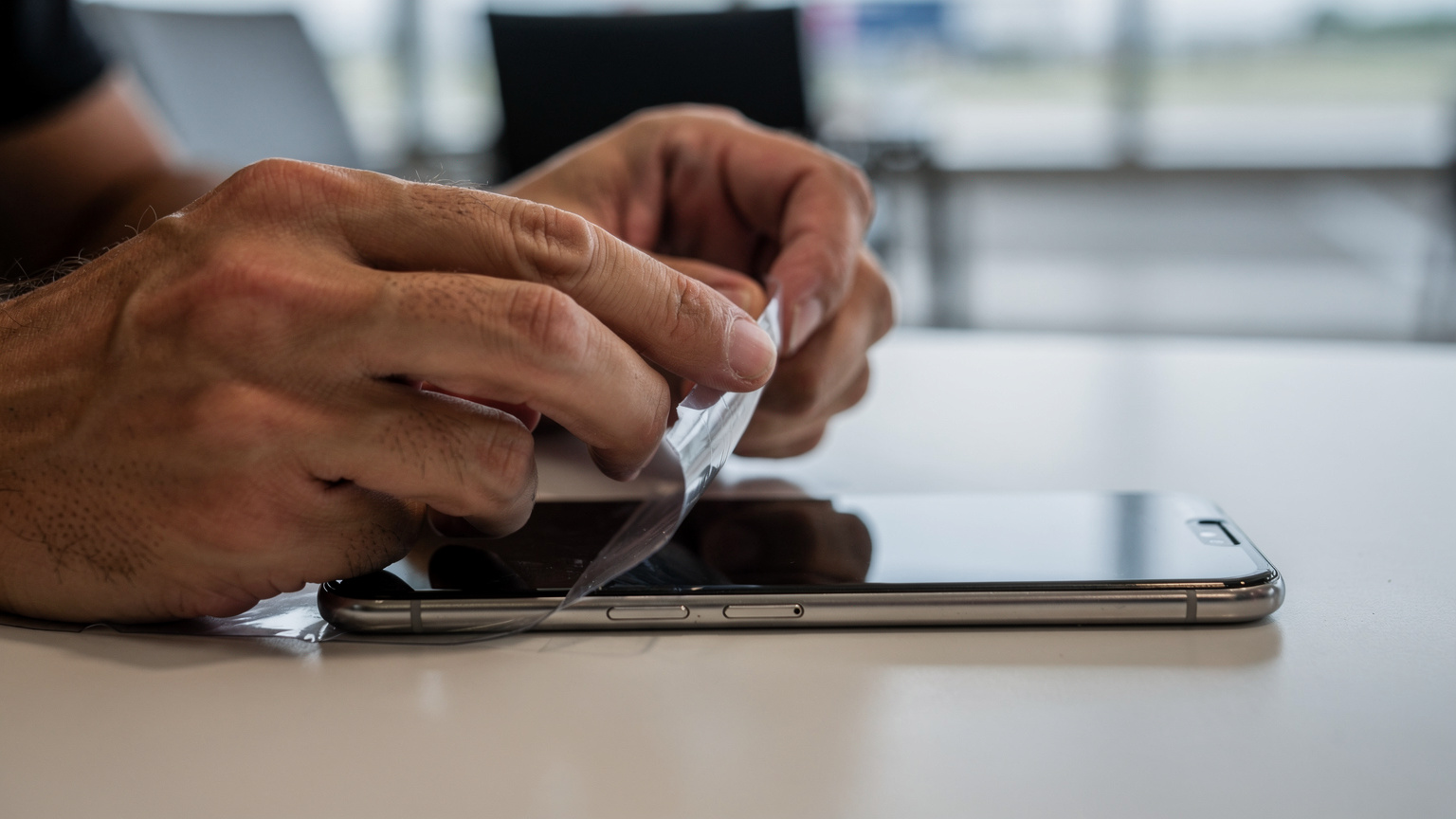 Bir kişinin elleri akıllı telefon ekranından koruyucu bir plastik filmi dikkatlice soyuyor ve taze bir ekranı ortaya çıkarıyor. Arka plan hafifçe bulanık, bir havaalanı salonu ortamını andırıyor.